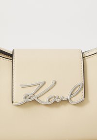 Beige leather handbag with a smooth texture, featuring a silver signature logo on the flap and a contrasting black zipper detail.