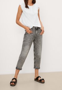 Mujer vestida con una blusa blanca sin mangas con hombros volantes, pantalones vaqueros grises remangados y sandalias negras con hebilla, de pie sobre un suelo beige.