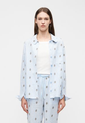 Frau trägt ein hellblau-weiß gestreiftes Hemd und eine Hose mit Bienendrucken über einem weißen Oberteil und steht vor einem schlichten weißen Hintergrund.