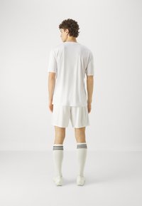 Weißes Sporttrikot mit kurzen Ärmeln, kombiniert mit weißen Shorts und knielangen Socken mit schwarzen Streifen. Minimalistisches Design, glatte Textur.