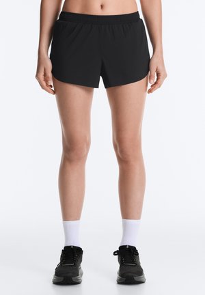 Femeie purtând pantaloni scurți sport negri, șosete albe până la gambă și pantofi de alergare negri, prezentată de la mijlocul trunchiului până la picioare pe fundal simplu.