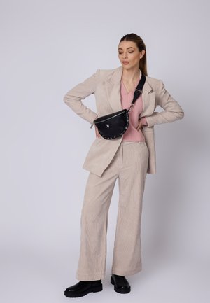 Černá kožená taška s zakřiveným tvarem a hřebíkovými akcenty, nošená přes rameno. Model v béžovém kordovém obleku a růžovém topu, s černými botami.