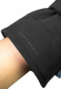 Schwarze Softshell-Stoffhülle mit glatter Textur, versehen mit genähten Nähten und einem "SOFTSHELL"-Label. Nahaufnahme von einem menschlichen Arm.