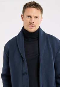 Homme portant un cardigan bleu marine à texture avec des boutons sur un pull à col roulé noir, regardant droit devant lui avec une expression neutre.