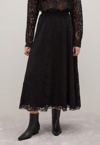 Falda midi de encaje negro con patrón floral, cintura elástica y textura transparente. Combinada con botas negras hasta el tobillo con puntera afilada.