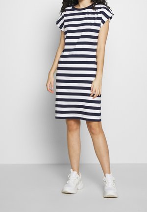 Žena v tmavě modrých a bílých proužkovaných šatech po kolena s krátkými rukávy a bílými teniskami stojí před jednobarevným pozadím.