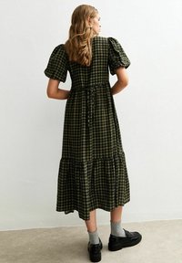 Robe maxi à carreaux verts avec manches bouffantes, taille cintrée et jupe à volants. Portée avec des chaussures noires et des chaussettes grises. Texture de tissu lisse.
