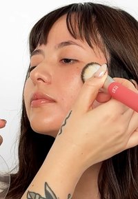 Pincel de maquillaje con mango rosa siendo utilizado en la mejilla de una persona, destacando una textura de piel suave e iluminada y sutiles detalles faciales.