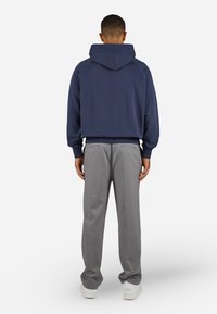 Man die wegkijkt, gekleed in een marineblauwe hoodie, grijze broek en witte sneakers, staand tegen een effen lichte achtergrond.