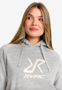 Grå hoodie med en framficka i kängurustil, dragskohuva och stor krämfärgad RVRC-logotyp på bröstet, tillverkad av mjukt bomullsblandat tyg.