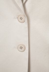 Detail blízkého pohledu na béžovou látkovou bundu se dvěma kulatými, neutrálními knoflíky a jemným prošíváním na levé straně.