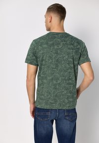 Zielona koszulka z krótkim rękawem z teksturowanym, abstrakcyjnym wzorem. Krój z okrągłym dekoltem, zestawiona z niebieskimi dżinsami.