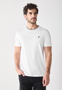 Weißes Baumwoll-T-Shirt mit kurzen Ärmeln, schwarzem Besatz am Kragen und einem kleinen schwarzen Vogel-Logo auf der linken Brust.