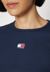Marinblå ribbad t-shirt med en broderad "TOMMY JEANS"-logotyp i en fyrkantig patch i röd, vit och blå färg.