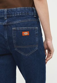 Κοντινό πλάνο ενός ατόμου που φοράει σκουρόχρωμα μπλε τζιν και καφέ μπλουζάκι, παρουσιάζοντας την πίσω τσέπη με μια μικρή πορτοκαλί ετικέτα Dickies.