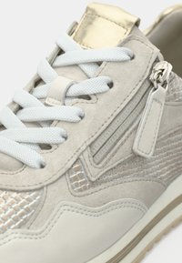 Αθλητικό παπούτσι σε ανοιχτό μπεζ χρώμα με υφασμάτινα πάνελ με υφή, λευκά κορδόνια και χρυσή λεπτομέρεια στη γλώσσα. Διαθέτει φερμουάρ και επενδεδυμένο κολάρο.