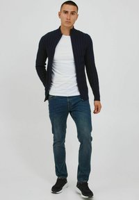 Mørkeblå strikket jakke med lodrette linjer, høj krave og lynlås lukning, parret med en hvid T-shirt, blå jeans og sorte sportsko.
