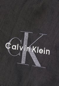 Svart tyg med broderad "Calvin Klein"-logotyp i vitt och grått. Texturen är slät med en subtil lyster.
