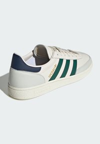 Białe skórzane sneakersy z granatowym zapiętkiem, zielonymi paskami i złotym logo. Posiadają teksturowaną podeszwę i płaskie sznurówki. Klasyczny design.