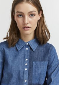 Camicia di jeans con colletto classico, bottoni frontali e tasca sul lato sinistro. Il tessuto presenta diverse sfumature di blu.