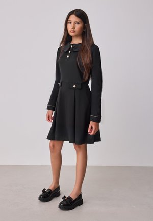 Jeune femme debout, portant une robe noire à manches longues avec des boutons en perle et des mocassins noirs avec des nœuds, fond neutre.
