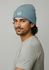 Light blue ribbed beanie with a square patch featuring a white mountain and wave design. Worn by a person in a gray shirt.