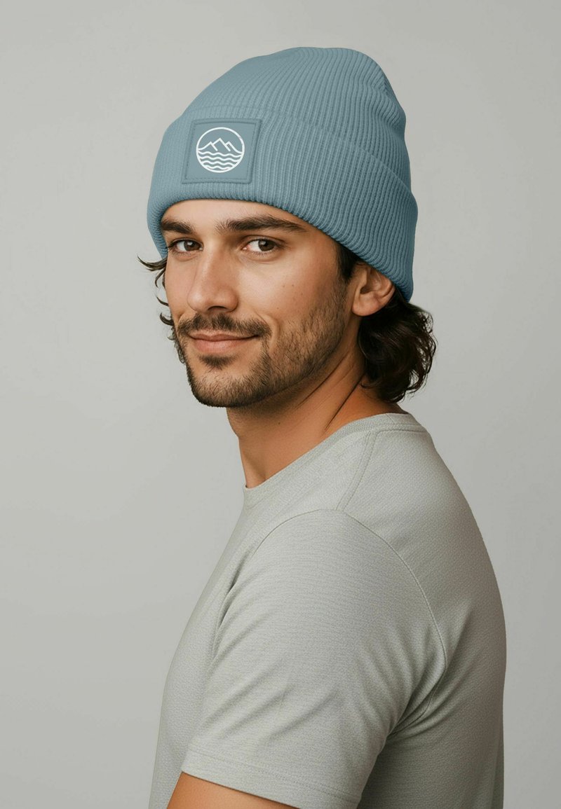 Light blue ribbed beanie with a square patch featuring a white mountain and wave design. Worn by a person in a gray shirt.