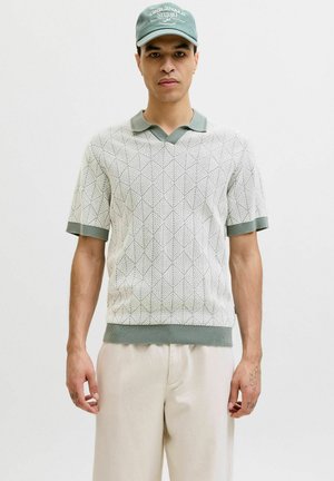 Junger Mann trägt ein gemustertes kurzärmeliges Strick-Poloshirt, beige Hose und eine grüne Kappe mit weißem Text, stehend vor einem einfarbigen Hintergrund.