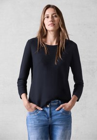 Maglietta nera textured a maniche lunghe con scollatura rotonda, abbinata a jeans azzurri chiari, mani in tasca, sullo sfondo neutro.
