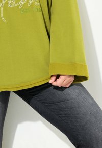 Sweatshirt vert à manches longues et texture douce, avec un texte brodé et un ourlet sombre contrastant, associé à un jean noir ajusté.
