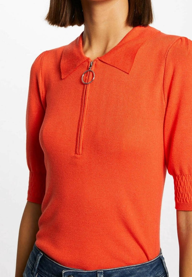 Femme portant un haut en maille orange vif à manches courtes avec col et fermeture éclair frontale dotée d’un anneau de tirage circulaire, associé à un jean bleu.