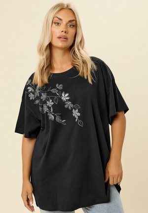 Blonde Frau trägt ein lockeres schwarzes T-Shirt mit silbernem Blumendesign auf der linken Schulter und hellblaue Jeans vor beigem Hintergrund.