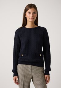 Pull en maille torsadée bleu marine avec des poignets et un ourlet côtelés. Il présente deux poches avant avec des accents de boutons dorés. Associé à un pantalon à carreaux.
