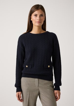 Marineblauer Kabelstrickpullover mit gerippten Bündchen und Saum. Verfügt über zwei Fronttaschen mit goldfarbenen Knopf-Akzenten. Kombiniert mit karierten Hosen.
