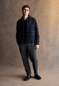 Ein junger Mann trägt eine dunkelblaue strukturierte Jacke, ein schwarzes Hemd, graue Hosen und schwarze Stiefel und steht vor einem neutralen braunen Hintergrund.