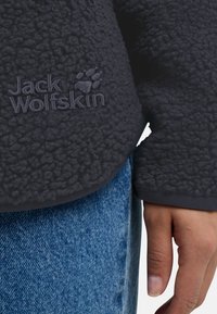 Charcoal fleece jack met een gestructureerd oppervlak, voorzien van een subtiele logo en ribgebreide manchetten, gecombineerd met denimstof die zichtbaar is bij de taille.