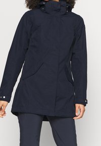 Abrigo impermeable de color azul marino con un cuello alto, bolsillos frontales y puños con botones a presión. Presenta un dobladillo recto y una textura suave.