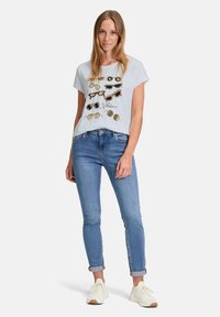 T-shirt bleu clair à manches courtes ornée de diverses illustrations de lunettes de soleil dans des couleurs contrastées, assortie à un jean bleu slim et des baskets blanches.