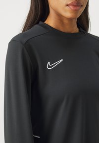 Černé dlouhé tričko Nike s kulatým výstřihem, na kterém je bílé vyšívané logo na hrudi a hladká, elastická textilie.