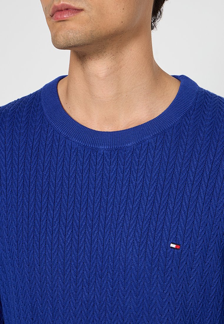 Blauwe gebreide trui met een gestructureerd, geribbeld patroon en een ronde hals. Beschikt over een klein geborduurd logo op de borst.