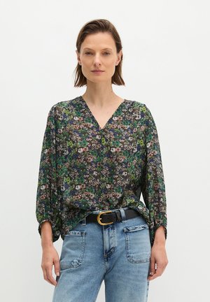 Frau trägt eine dunkle, florale V-Ausschnitt-Bluse mit dreiviertel Ärmeln, in hellblaue Jeans gesteckt, dazu schwarzer Gürtel mit goldener Schnalle.