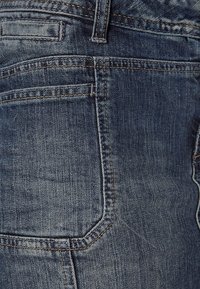 Vue en gros plan de la zone de la poche arrière d'un jean en denim bleu délavé avec des coutures visibles et des passants de ceinture.