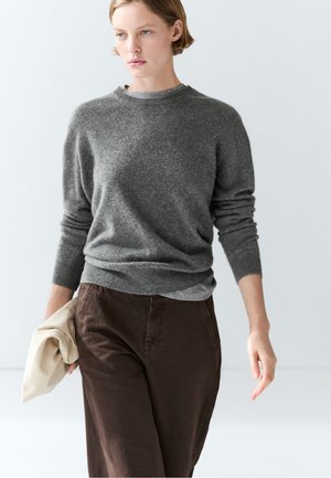 Ung person iført en grå sweater og brune bukser, der holder et beige klæde, gående mod en ensfarvet lys baggrund.
