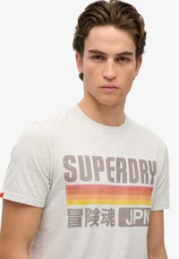 Camiseta de algodón gris claro con un atrevido logo "SUPERDRY" en tonos de gris oscuro y rayas en degradado de rojo anaranjado; mangas cortas y escote redondo.