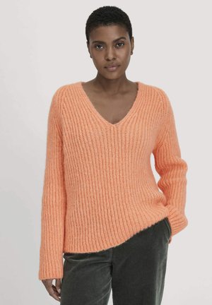Vrouw met kort haar, gekleed in een perzikkleurige ribgebreide V-hals trui en donkergroene fluwelen broek, poseert met één hand in haar zak.