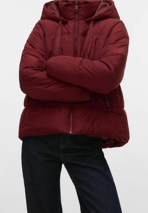Veste matelassée bordeaux avec capuche, fermeture éclair à l'avant et poches, portée avec un jean foncé, bras croisés sur la poitrine.