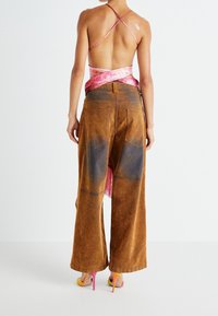 Zadní pohled na modelku, která má na sobě růžový saténový překládaný top a hnědé široké kalhoty s vybledlými oblastmi, doplněné jasnými lodičkami na vysokém podpatku.