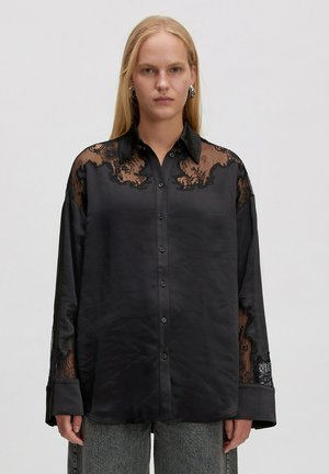 Schwarze Satinbluse mit Spitzenakzenten an den Schultern und Ärmeln, mit durchgehender Knopfleiste und langen Ärmeln. Strukturierte Spitzenverzierung.