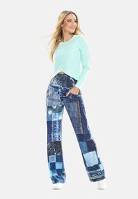Helle türkisfarbene Langarmbluse mit überkreuztem Saum; blaue Jeanshose mit Patchwork-Design in verschiedenen Texturen und Blautönen.