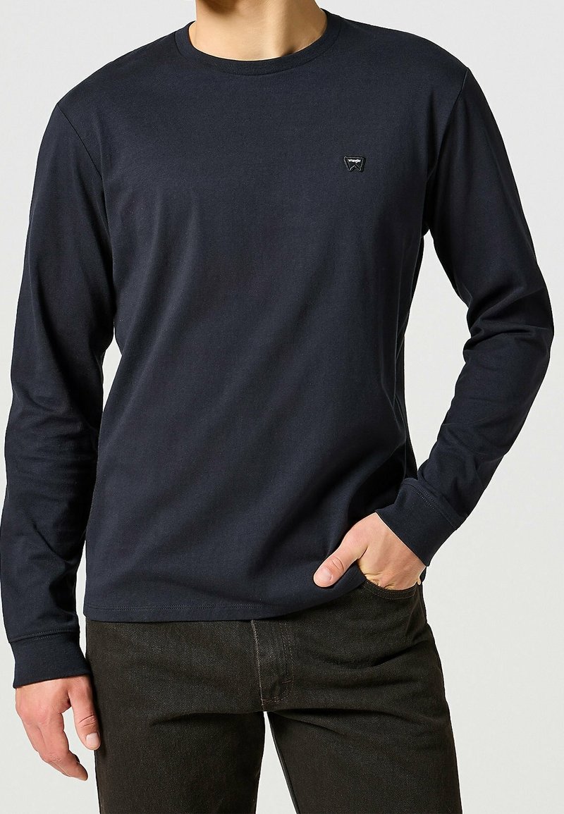 Hombre vistiendo una camiseta de manga larga negra tipo crewneck con un pequeño logo en el pecho y pantalones oscuros, con una mano en el bolsillo.
